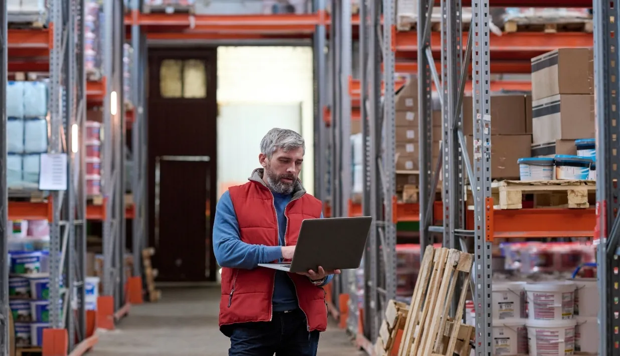 Warehouse worker reviewing an item and operating context on a laptop between storage racks